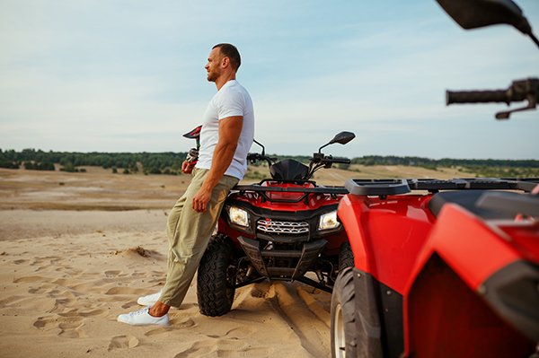 Brutal man poses near atv in desert sands. Male person on quad bike, sandy race on quadbike, dune safari in hot sunny day, 4x4 extreme adventure, quad-biking